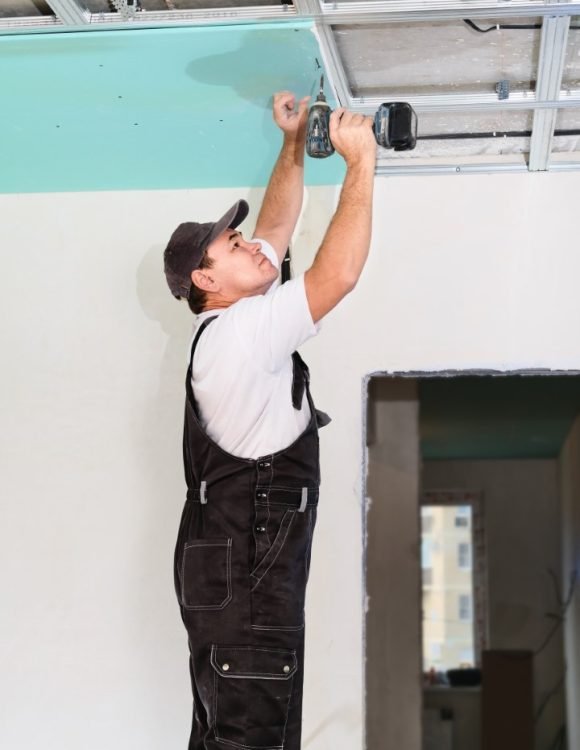 Worker assembles a suspended ceiling with plasterboard and attaches the plasterboard to the metal frame of the ceiling using an electric screwdriver.